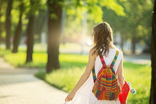 Portrait of lovely urban girl in white dress with a pink skateboard. Happy smiling woman. Girl holding a plastic skate board outdoors. City life. Back view