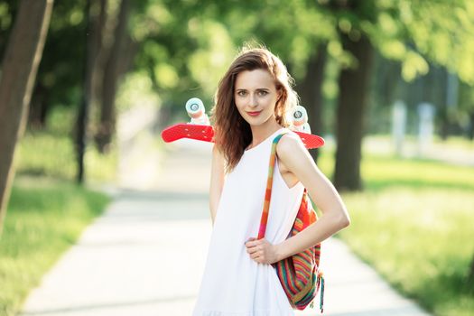 Portrait of lovely urban girl in white dress with a pink skateboard. Happy smiling woman. Girl holding a plastic skate board outdoors. City life.