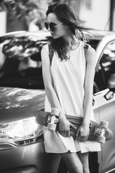 Portrait of lovely urban girl in white dress with a skateboard. Happy smiling woman. Pretty girl is standing near a car. Black and white photography.