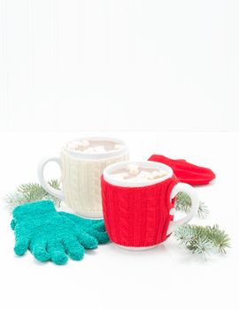Two mugs pf creamy hot chocolate with mittens and spruce branches.