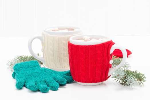 Two mugs of creamy hot chocolate with gloves and spruce.