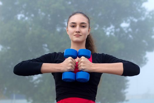 Young pretty slim fitness sporty woman exercises with weights dumbells during training workout outdoor