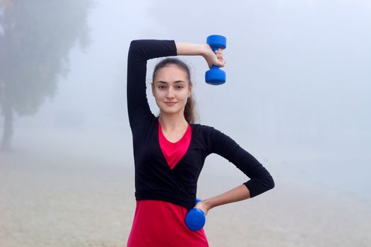 Young pretty slim fitness sporty woman exercises with weights dumbells during training workout outdoor