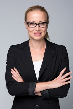 Portrait of beautiful young caucasian woman in business attire wearin black eyeglasses standing with arms crossed against gray background.