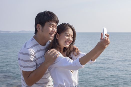 portrait of younger asian man and woman taking a photograph by smart phone ,relaxing vacation time at sea side happiness emotion