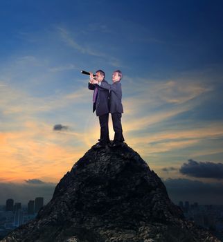 two of asian business man standing on top of rock cliff and looking by spying lens with skyscraper background