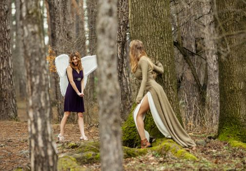 Two beautiful women in a fairytale forest. One as fairy with wings , the other is looking at her through the trees.