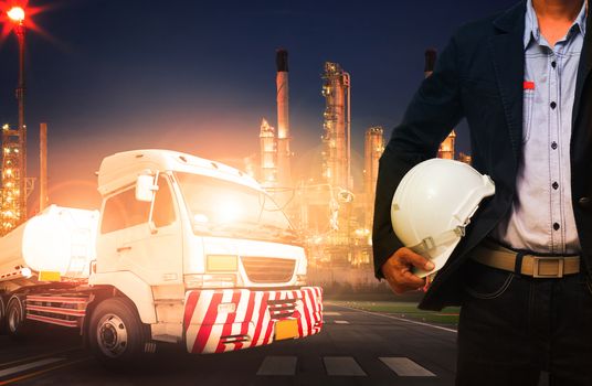 engineer holding hard hat standing against beautiful lighting of oil refinery in heavy petrochemical industry and big oil container truck use for petroleum industrial and land transport business