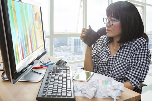 working woman with hot beverage cup in hand looking to computer monitor on working table crumpled paper sheet on deck