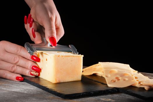 Female hands with beautiful bright manicure cut cheese whetstone by means of a cheese slicer on a black slate plate in style a rustic