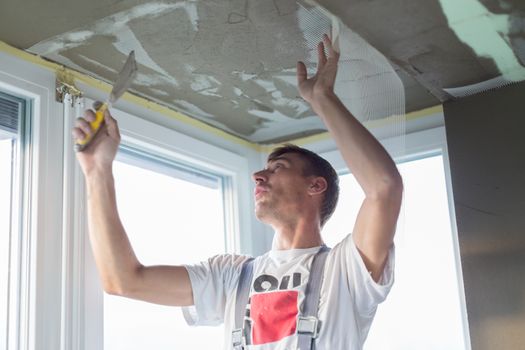 Thirty years old manual worker with wall plastering tools renovating house. Plasterer renovating indoor walls and ceilings with float and plaster. Wall mash installation. Construction finishing works.