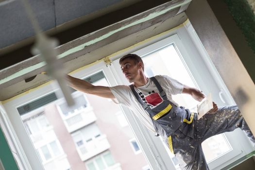 Thirty years old manual worker with wall plastering tools renovating house. Plasterer renovating indoor walls and ceilings with float and plaster. Wall mash installation. Construction finishing works.