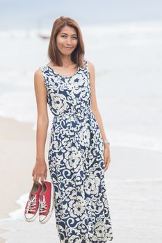 young asian woman with smiling face wearing long dress standing with sneaker in hand on sea beach