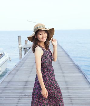 portrait of beautiful young woman wearing wide straw hat and long dress standing with happiness emotion on piers at sea beach 