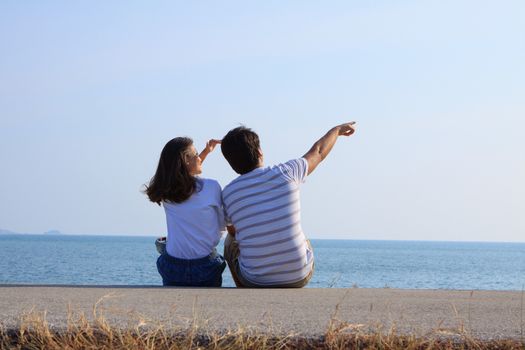 couples of man and woman sitting at sea side and point and looking to blue sky 