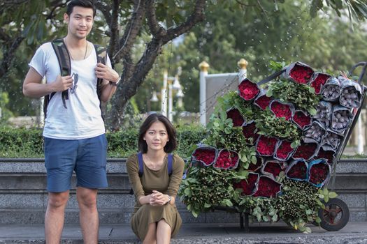 portrait couples of younger asian traveling man and woman sitting at road side beside red roses flower bouquet