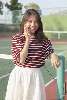 portrait of young beautiful asian woman talking smartphone in tennis course sport field use for modern life and digital telecommunication technology