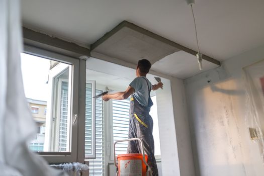 Thirty years old manual worker with wall plastering tools renovating house. Plasterer renovating indoor walls and ceilings with float and plaster. Wall mash installation. Construction finishing works.