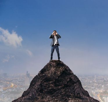 young business man standing on top of mountain and spying by binocular looking for something