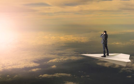 business man standing and spying binocular on papet plane against sun rising over cloud scape