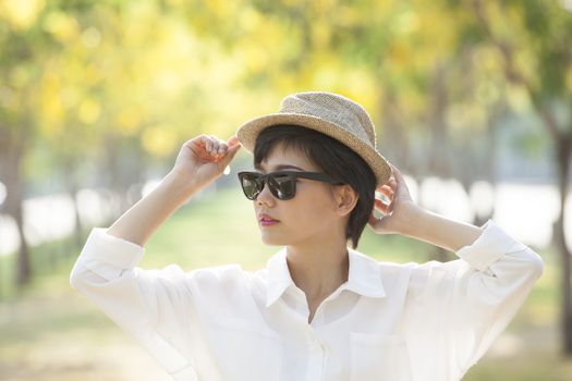 portrait of young beautiful asian woman wearing sun glasses and straw hat against yellow flowers blooming in park 