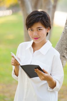 portrait of young beautiful woman wearing white shirt standing in park with book in hand looking to camera and smiling face with happiness emotion