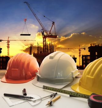 engineer working table plan, home model and writing tool equipment against building construction crane with evening dusky sky 