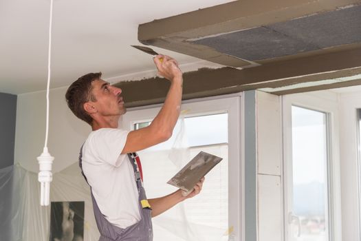 Construction worker wearing worker overall with wall plastering tools renovating apartment house. Plasterer renovating indoor walls and ceilings with float and plaster. Construction finishing works.