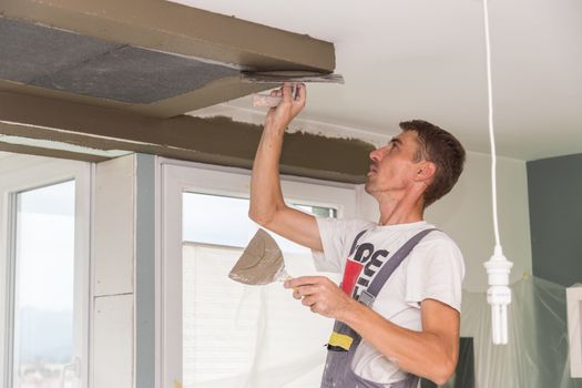 Construction worker wearing worker overall with wall plastering tools renovating apartment house. Plasterer renovating indoor walls and ceilings with float and plaster. Construction finishing works.