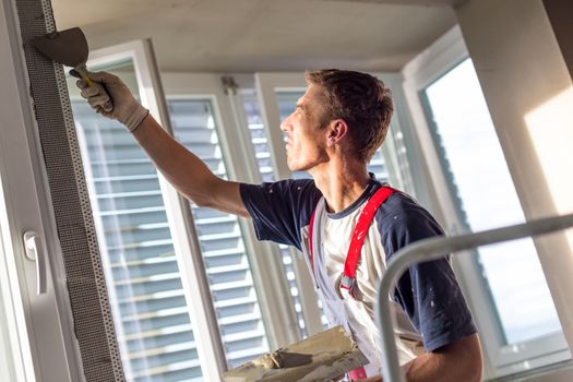 Construction worker wearing worker overall with wall plastering tools renovating apartment house. Plasterer renovating indoor walls and ceilings with float and plaster. Construction finishing works.