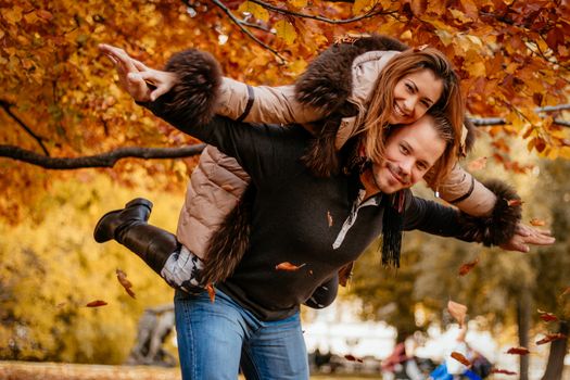 Beautiful young couple enjoying a piggyback in sunny park in autumn colors. Looking at camera.