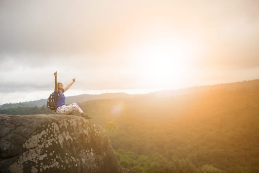 backpacker woman relaxing and victory hand rising on rock cliff and sun set sky above