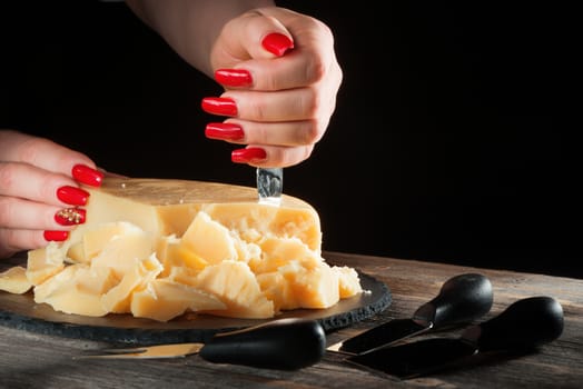 Female hands with bright beautiful manicure break away pieces of hard cheese like parmesan by means of a special knife on a plate from black slate in style a rustic.