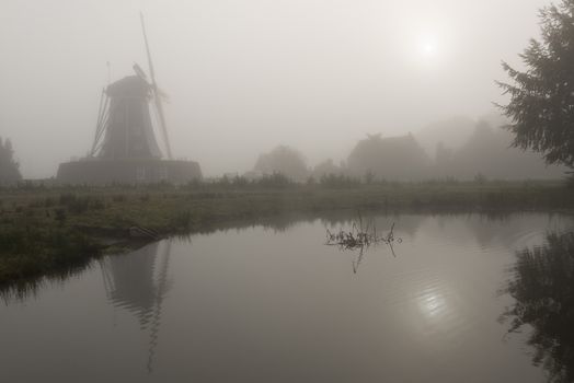 Historic corn mill called The Bataaf in Winterswijk in the early morning fog with an emerging Sun that reflectes in the nearby pond

