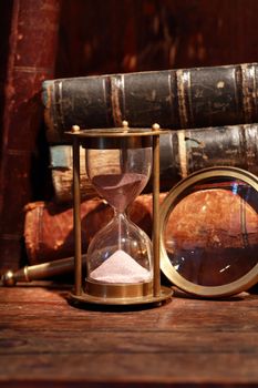 Still life with vintage hourglass near magnifying glass against old books