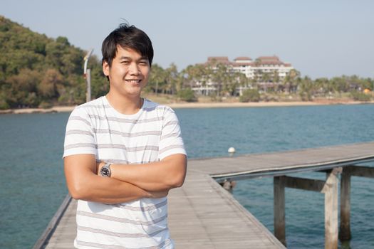 portrait of asian smart man standing and smiling with happy face on wood piers beside blue sea and beach