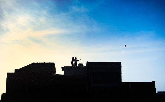 Two boys on the rooftop play with kites. A silhouette against the background of the blue sky with light clouds. 