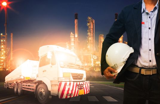 engineer holding hard hat standing against beautiful lighting of oil refinery in heavy petrochemical industry and big oil container truck use for petroleum industrial and land transport business