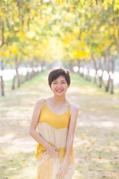 portrait of young beautiful asian woman wearing yellow long dress with smiling face ,happiness emotion standing in flowers blooming park vertical form