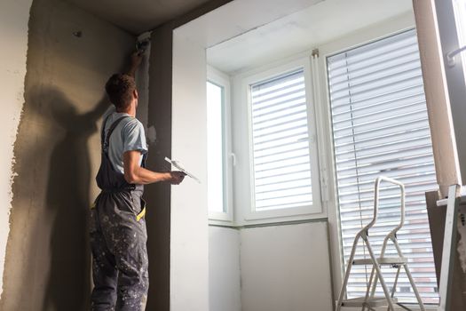Construction worker wearing worker overall with wall plastering tools renovating apartment house. Plasterer renovating indoor walls and ceilings with float and plaster. Construction finishing works.