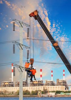electrician worker working on high voltage electric pole with crane against factory building background