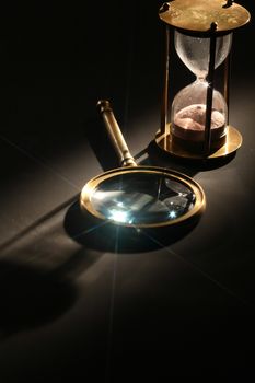 Vintage hourglass near magnifying glass on dark background with long shadow