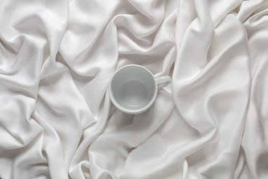 White ceramic empty coffee cup on the silk white fabric. Top view