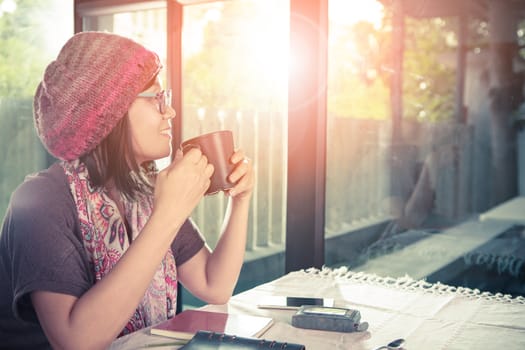 asian younger woman and hot coffee mug in hand smiling with happiness emotion sitting beside mirror window against beautiful sun light ,process warming color mood and tone