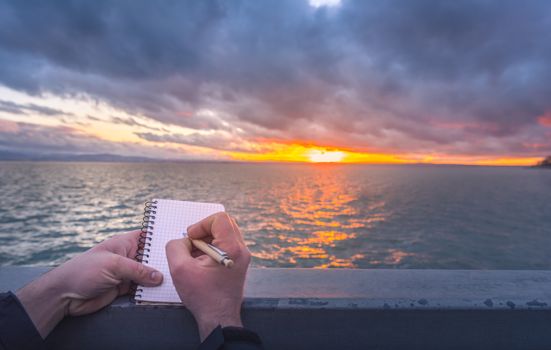 Conceptual image with a man writing in a spiral notebook while the sun sets down over the Bodensee lake.