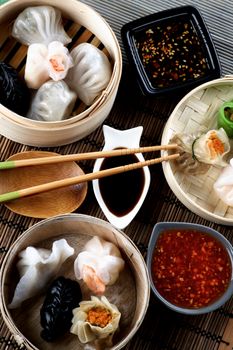 Arrangement of Various Dim Sum in Bamboo Steamed Bowls, Black and Red Chili Sauces, Soy Sauce and Chopsticks closeup on Straw Mat background. Top View