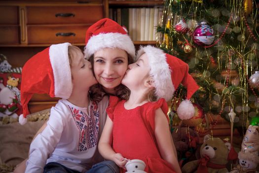 Two kids kissing their mother at cheek under Christmas tree