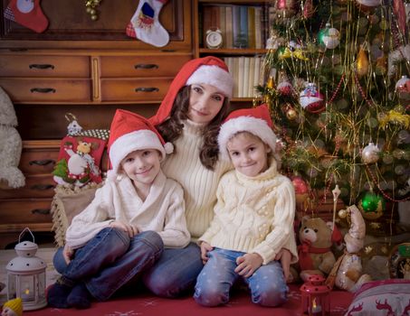 Two children sitting with mother under Christmas tree in hats