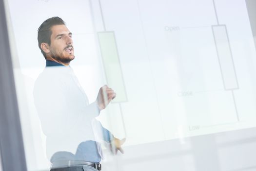 Business man making a presentation in front of whiteboard. Business executive delivering a presentation to his colleagues during meeting or in-house business training. View through glass.