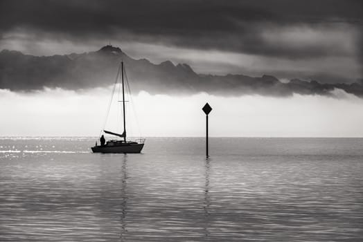 Black and white landscape with a boat sailing on the Bodensee lake, with mountains and morning mist at the horizon.